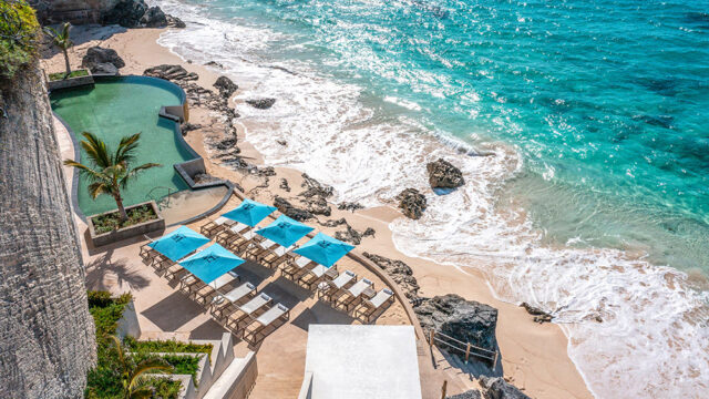Oceanfront infinity pool with lounge chairs and blue umbrellas on a sandy beach.