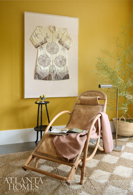 Rattan lounge chair with pink throw, book, and glasses. Yellow wall with framed robe.