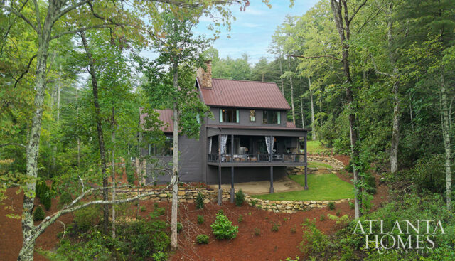 Dark gray house with brown metal roof and stone chimney, surrounded by trees.