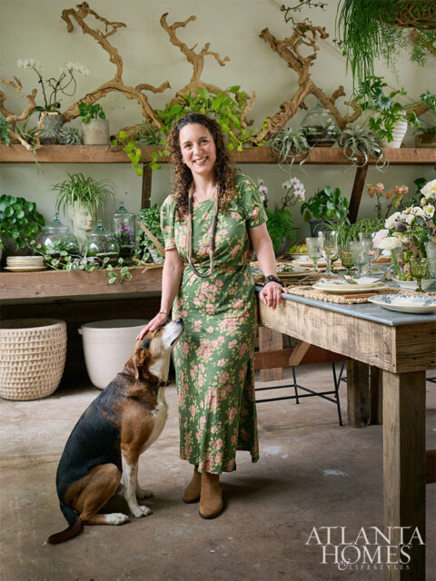 Woman in floral dress with dog, standing by a rustic table set for dining, with shelves of plants