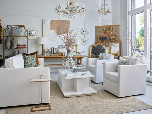 Living room with white sofas, chairs, and a white coffee table. Gold accents on mirrors and shelves.