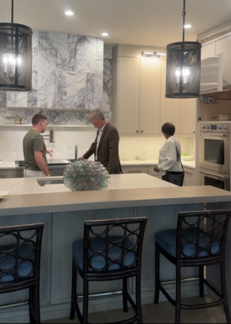 Kitchen with marble backsplash, gray island, and cream cabinetry. Three people are present.