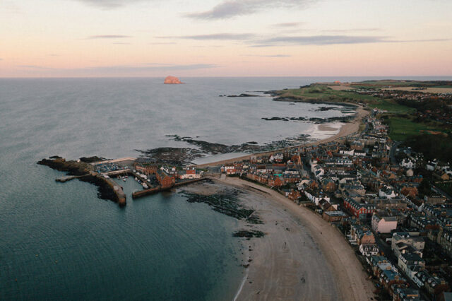 Coastal town with harbor and beach at sunset, with a small island offshore.