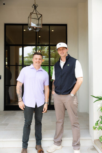 Two men standing on a porch. The man on the left wears a lavender shirt and dark jeans.