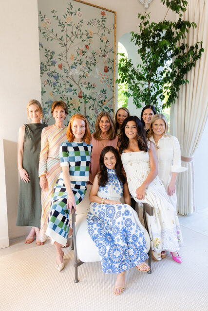 Group of eight women posing indoors, with a floral mural and large plant behind them.