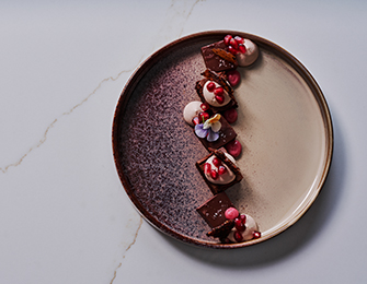 Dessert on a speckled plate with chocolate, cream, pomegranate seeds, and edible flowers.