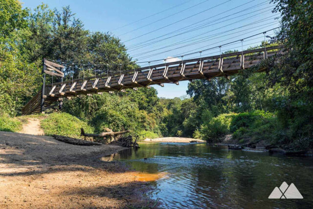 Wooden suspension bridge over a river surrounded by lush green trees under a clear blue sky.