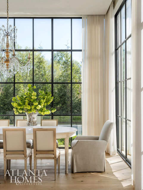 Dining room with oval table, upholstered chairs, crystal chandelier, and large windows.