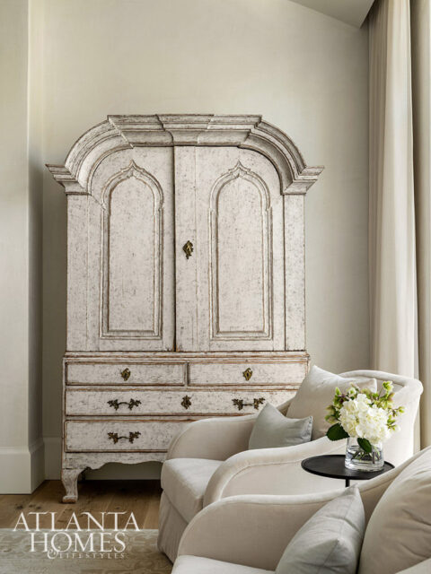Antique distressed white armoire with arched doors and drawers, flanked by upholstered chairs.