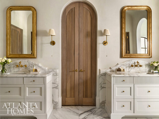 White vanities with marble counters flank a fluted wooden door. Gold mirrors hang above each vanity.