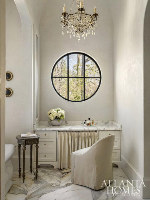 White bathroom with marble floors, circular window, crystal chandelier, and vanity