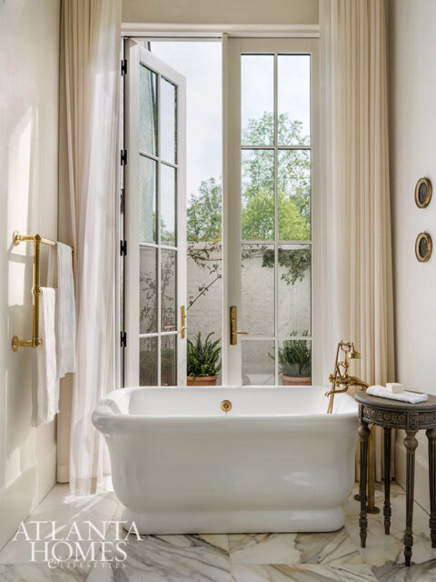 White freestanding bathtub with brass fixtures in a bathroom with marble floors and sheer curtains.