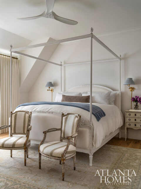 White canopy bed with blue throw, two striped armchairs, and a vintage nightstand.