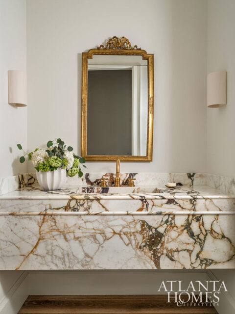 Bathroom with marble vanity, gold mirror, and brass fixtures. White flowers in vase.