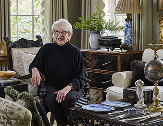 Elderly woman with white hair wearing glasses and a black sweater sits in an ornate chair in a