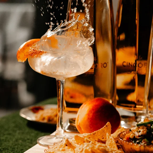 Cocktail with ice splash, peach slice, and chili rim garnish.