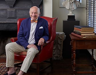 Man in a red armchair, wearing a blue blazer and bowtie.