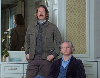 Two men in a bathroom, one wearing a jacket, the other in a suit.