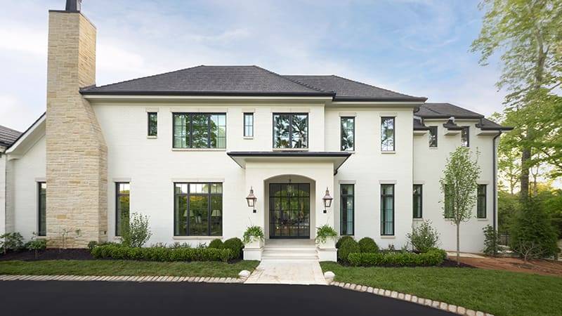 Two-story white brick home with stone chimney and black-framed windows, landscaped front yard.